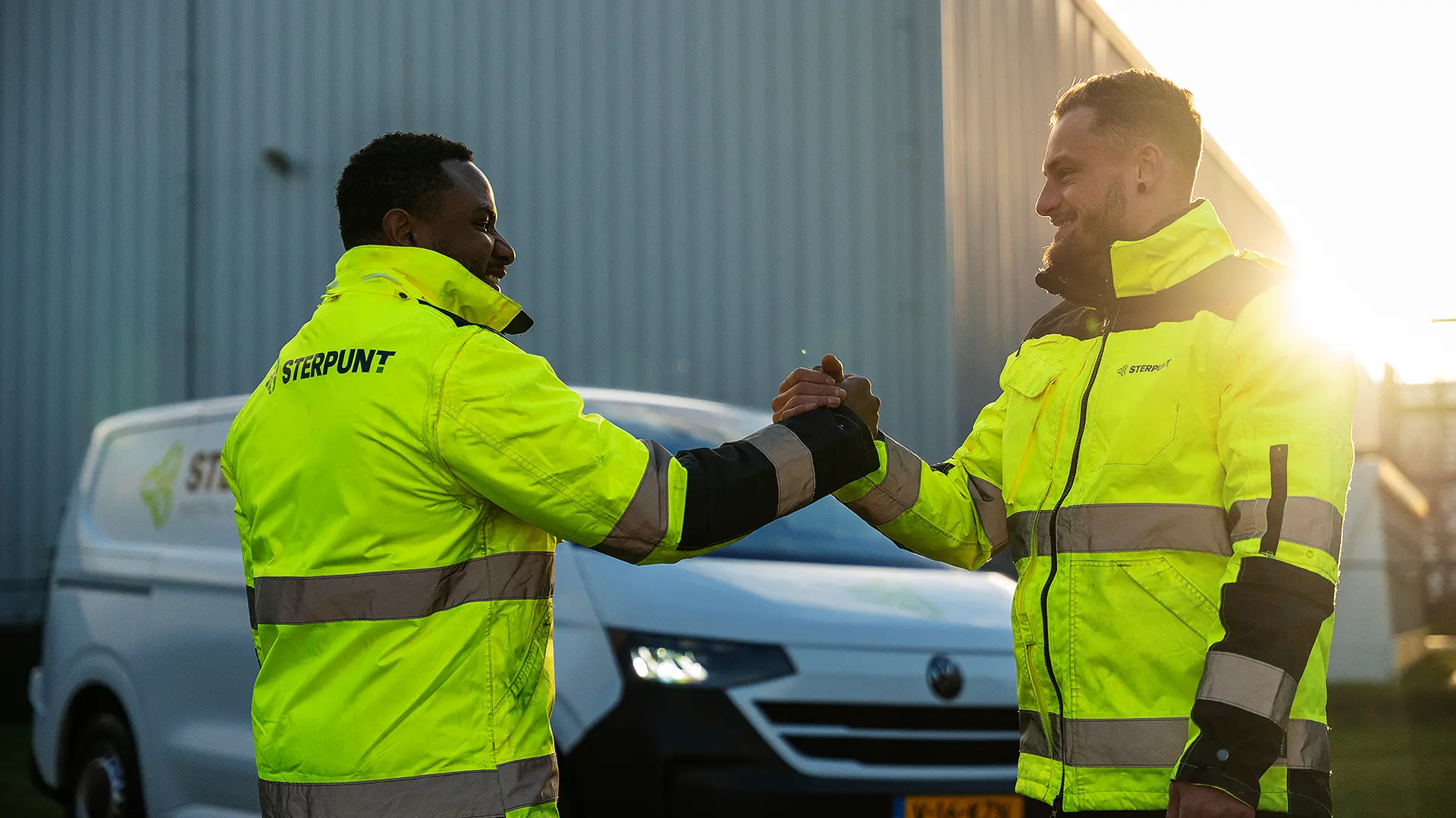 Twee monteurs in een neonkleurige jas van Sterpunt die elkaar de hand schudden met een werkbus op de achtergrond.